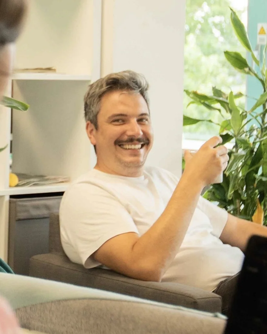 Anton Hannig lacht in einem Sessel, hinter ihm Zimmerpflanzen und ein Fenster